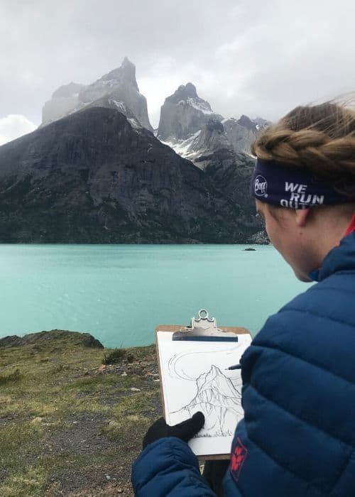Vicki Speck, una artista, está pintando frente a un lago con montañas al fondo.