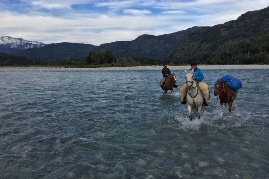 Cabalgata de dos personas montando a caballo por un río con montañas al fondo.