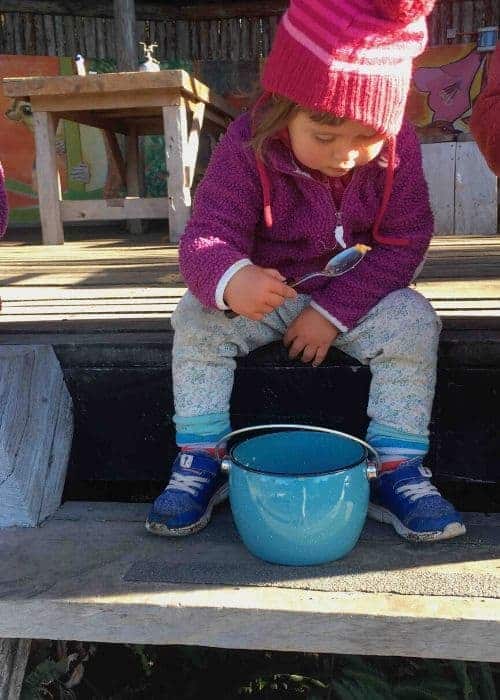 Una niña pequeña se sienta en un banco de madera y come de un cuenco azul mientras acampa.