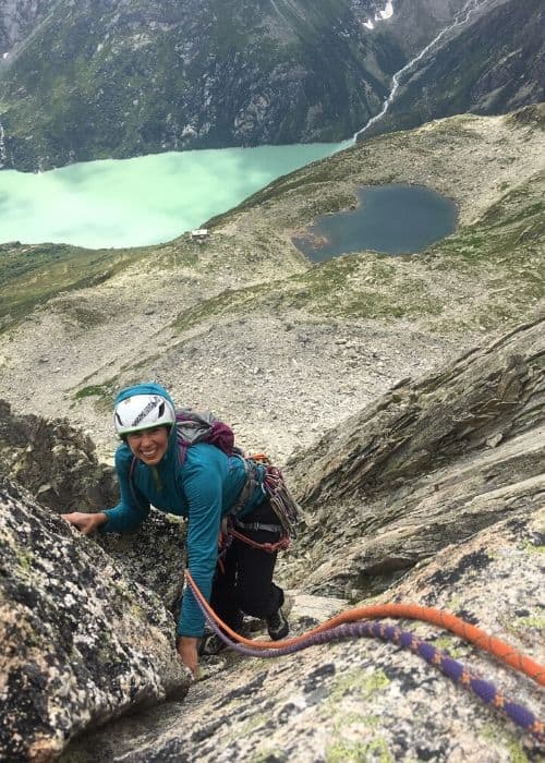 Una mujer practicando escalada mientras ascendía una montaña multilargo, con un impresionante lago como telón de fondo.