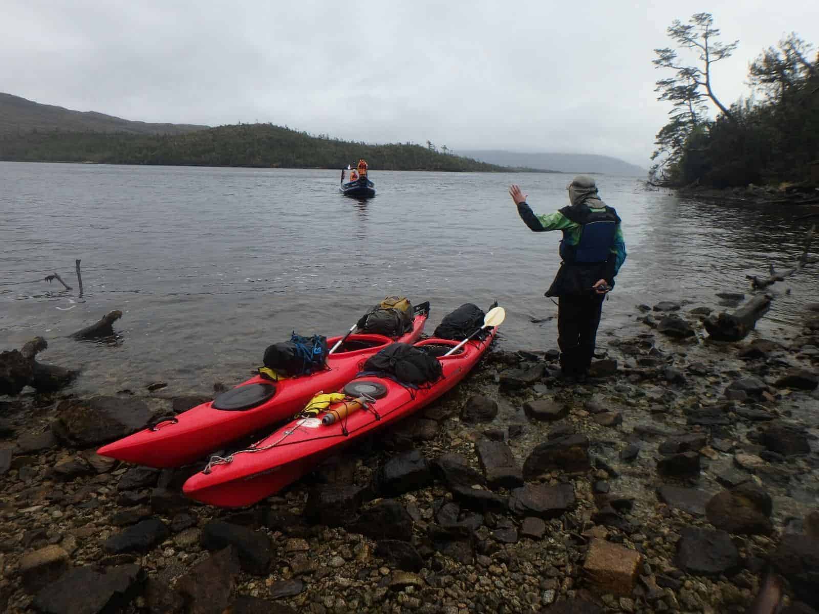 Una persona parada junto a dos kayaks en una orilla rocosa con vistas a un glaciar.