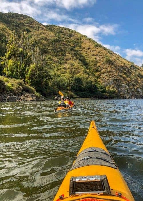 Dos personas acampan mientras navegan en kayak por un río con colinas al fondo.