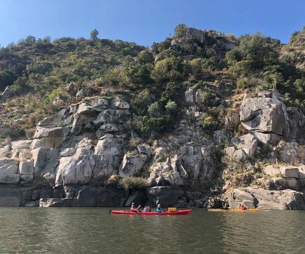 Un grupo de personas acampando y navegando en kayak por un río junto a un acantilado rocoso.