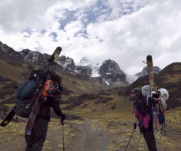 Dos personas paradas en un camino de tierra a gran altura con esquís en la espalda.