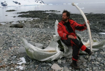 Cristián Donoso, un hombre con chaqueta roja, sentado en una roca junto a un iceberg en la Antártida.