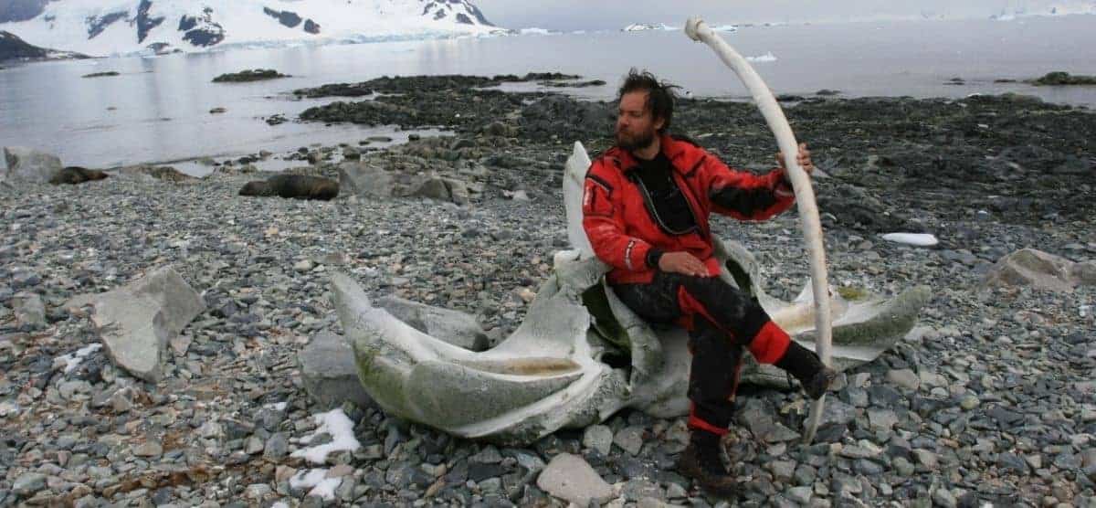 Cristián Donoso, un hombre con chaqueta roja, sentado en una roca junto a un iceberg en la Antártida.