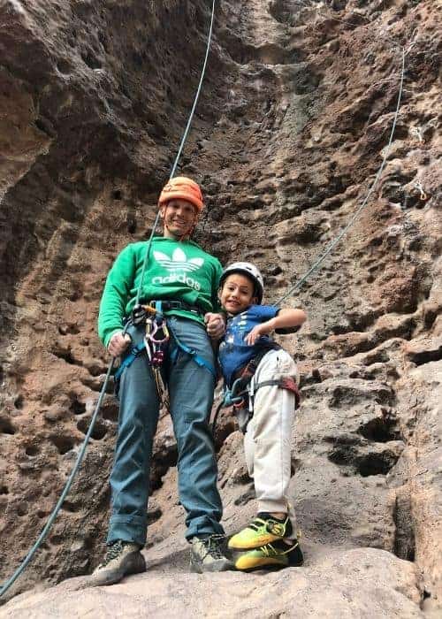 Un hombre y un niño abrazan la aventura mientras se paran frente a una pared de roca, liberándose de su zona de confort y creando preciados recuerdos familiares al aire libre.