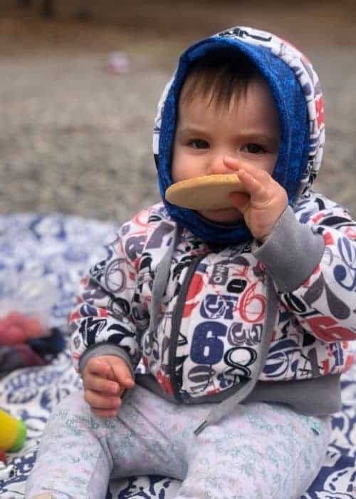 Un bebé disfruta de un picnic de invierno con su familia al aire libre, vistiendo una acogedora sudadera con capucha mientras come delicadamente un donut.