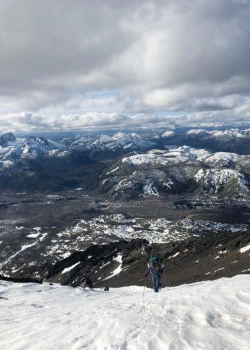 Una persona subiendo una montaña nevada con el majestuoso Volcán Lanín al fondo.