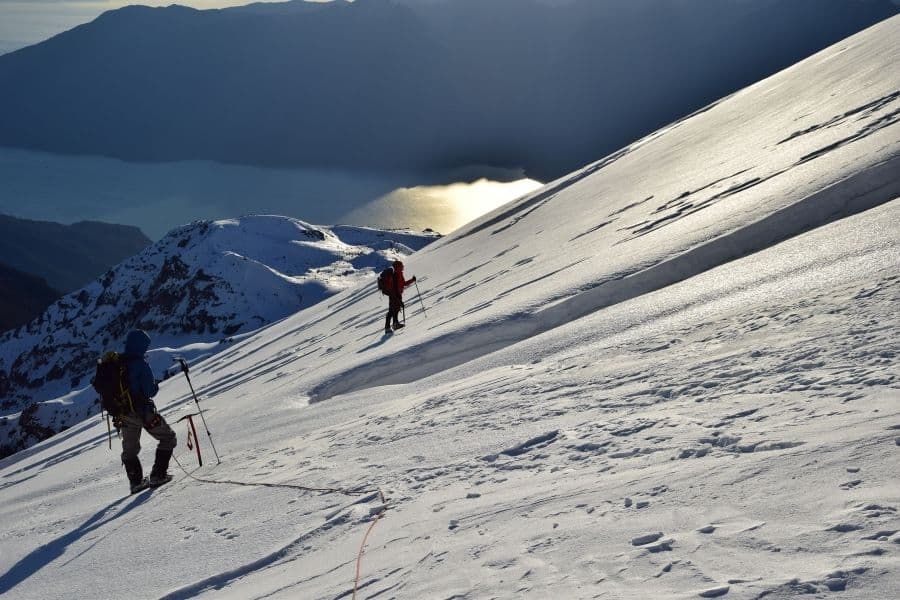 Dos personas subiendo una montaña nevada con un lago al fondo en los Andes.
