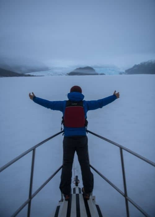 Un hombre parado en la cubierta de un barco con los brazos extendidos, disfrutando de las impresionantes vistas de Caleta María en Tierra del Fuego.