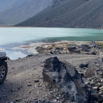 Un jeep Overland está estacionado en un camino rocoso cerca del Cajón Las Leñas, con vista a un lago.