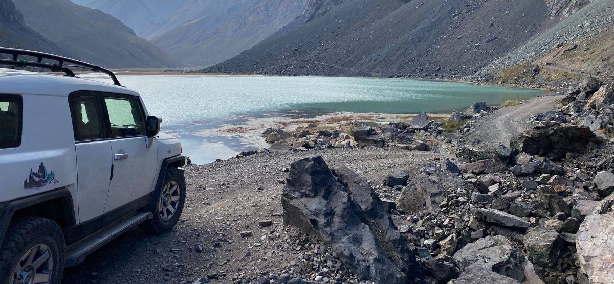 Un jeep Overland está estacionado en un camino rocoso cerca del Cajón Las Leñas, con vista a un lago.