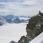 Un hombre parado en la cima de una montaña nevada, rodeado de agua y la amenaza del frío extremo.