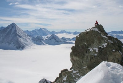 Un hombre parado en la cima de una montaña nevada, rodeado de agua y la amenaza del frío extremo.