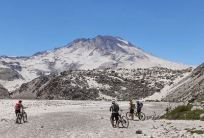Un grupo de personas descabezadas andando en bicicleta por una zona nevada con montañas volcánicas al fondo.