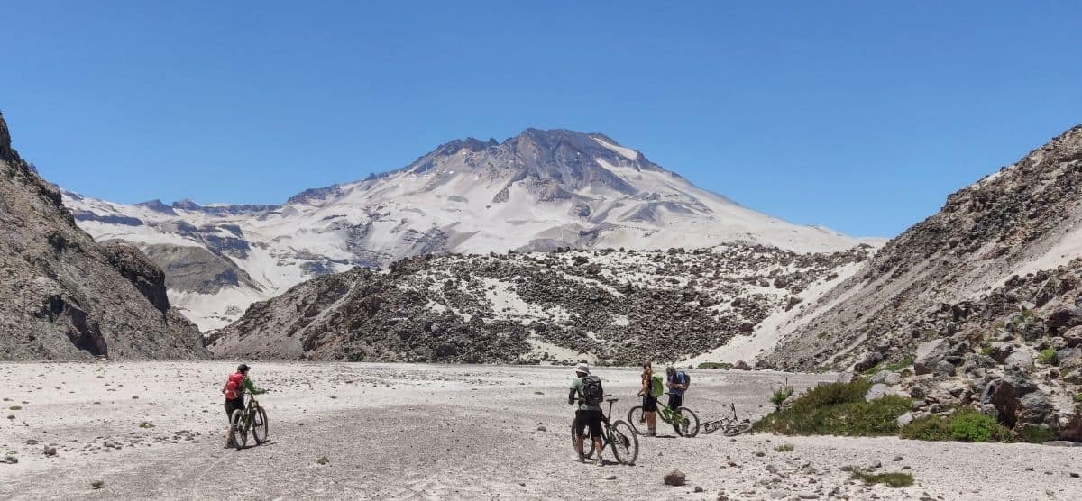 Un grupo de personas descabezadas andando en bicicleta por una zona nevada con montañas volcánicas al fondo.