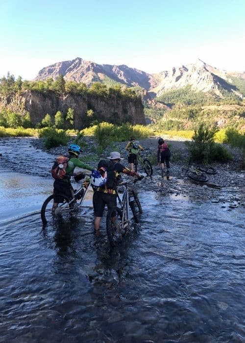 Un grupo de ciclistas de montaña descabezan un río usando sus bicicletas mientras disfrutan de la impresionante vista de un volcán cercano.