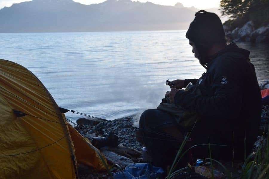 Un hombre sentado frente a una tienda de campaña cerca de una masa de agua en la Patagonia, contemplando su próximo ascenso a Primer Ascenso en La Muela.